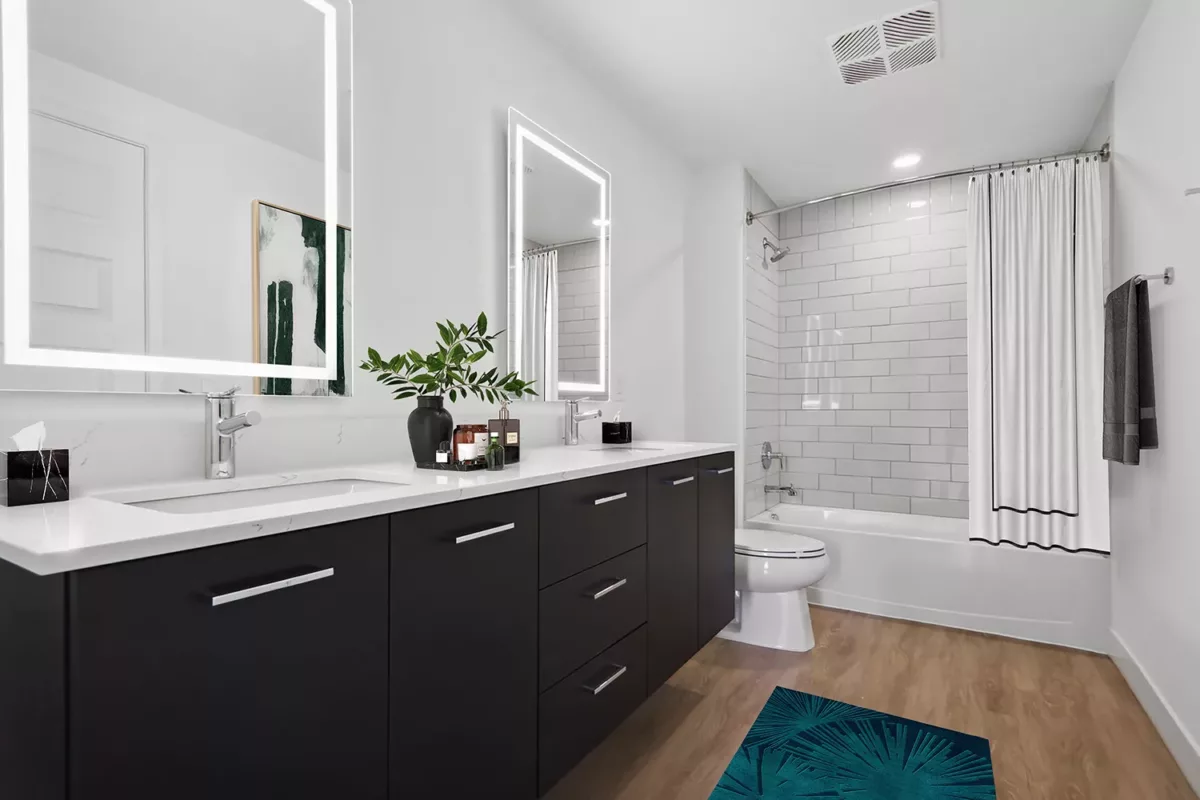 Modern bathroom with a black double-sink vanity, illuminated mirrors, white tiled shower, and greenery accents on the counter.