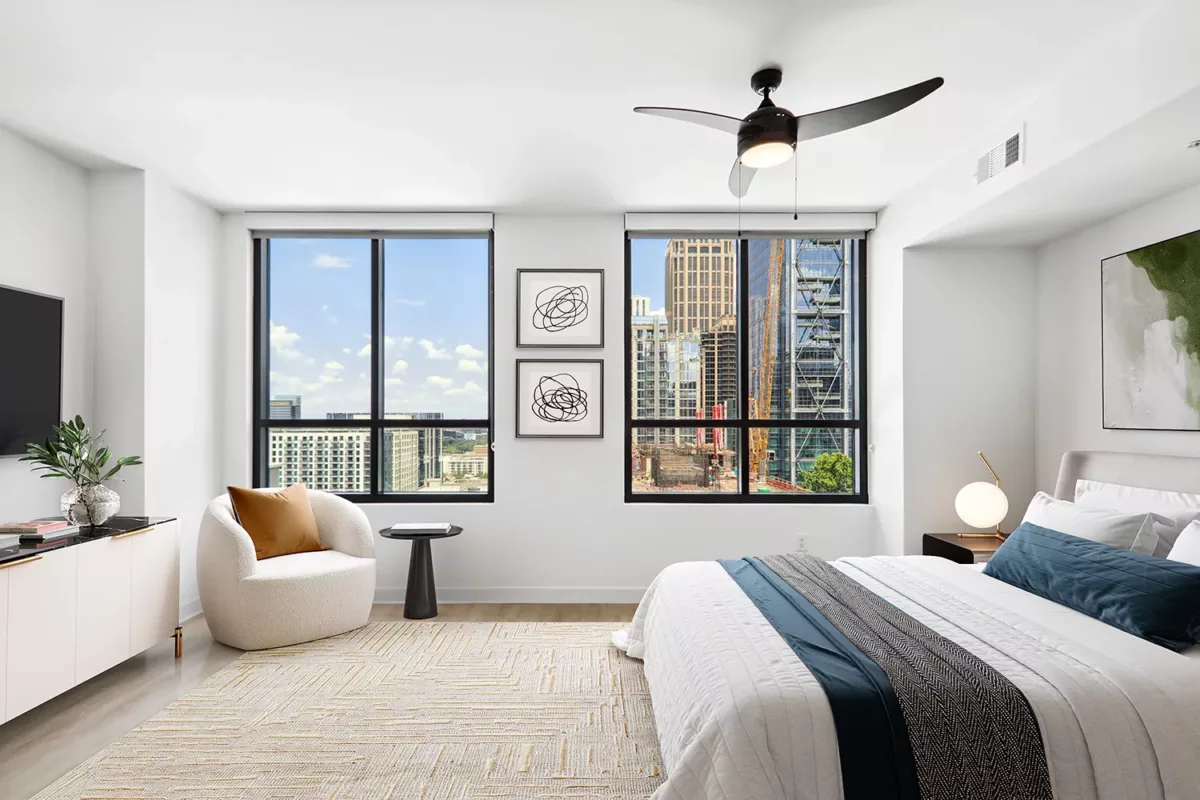 Bright bedroom with large windows showcasing a city skyline, modern furniture, and a ceiling fan.