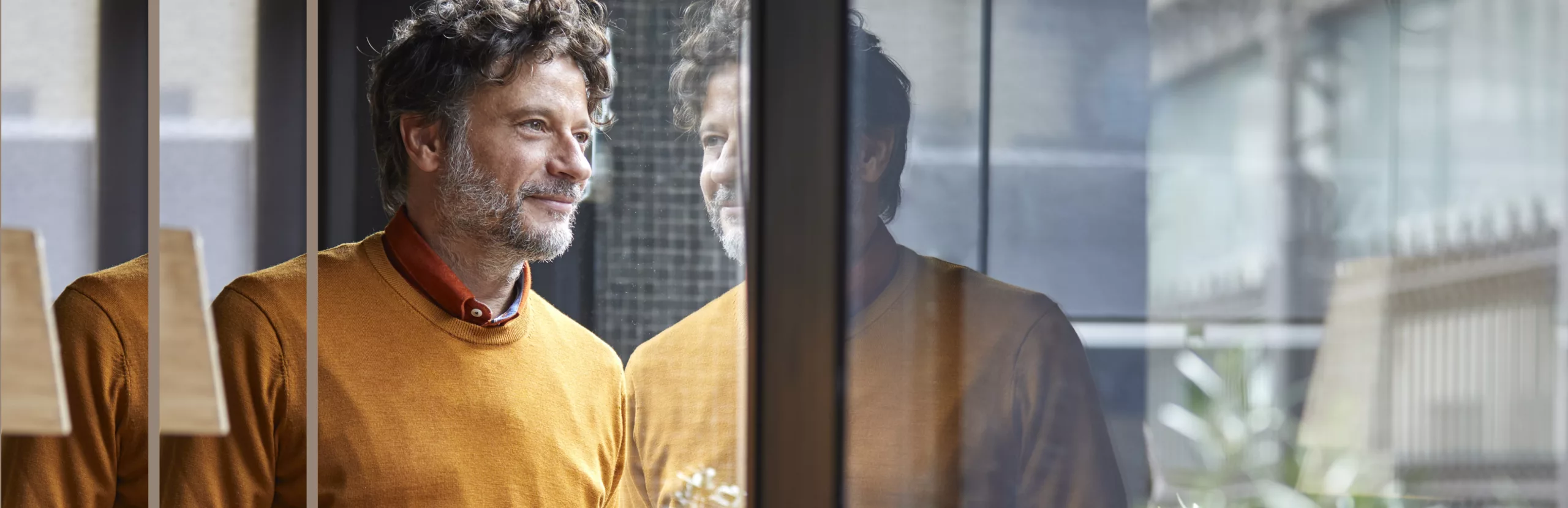 A man in a mustard yellow sweater and red collared shirt looking out a window with his reflection visible in the glass.