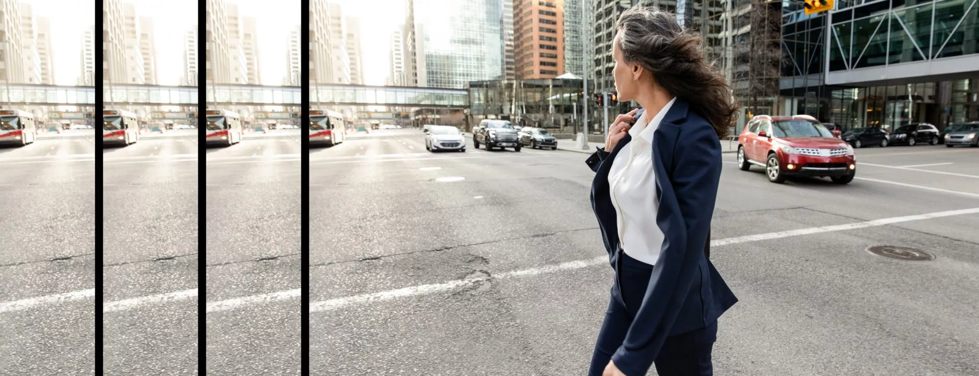 A woman in a business suit crossing a city street, with cars and tall buildings in the background. Her hair is blowing in the wind as she looks to the side.