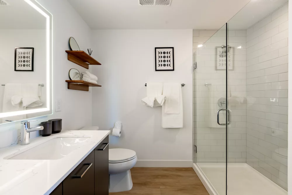 Bright bathroom featuring a glass-enclosed shower, lighted mirror, wooden shelves, and white towels on a rack.